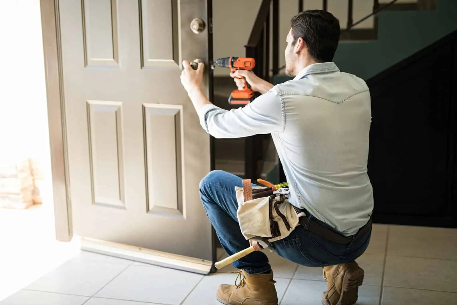 A man wearing a tool belt is kneeling by an open door, using a power drill to work on the entry doors lock. He is indoors, near a staircase, performing maintenance or repair work.