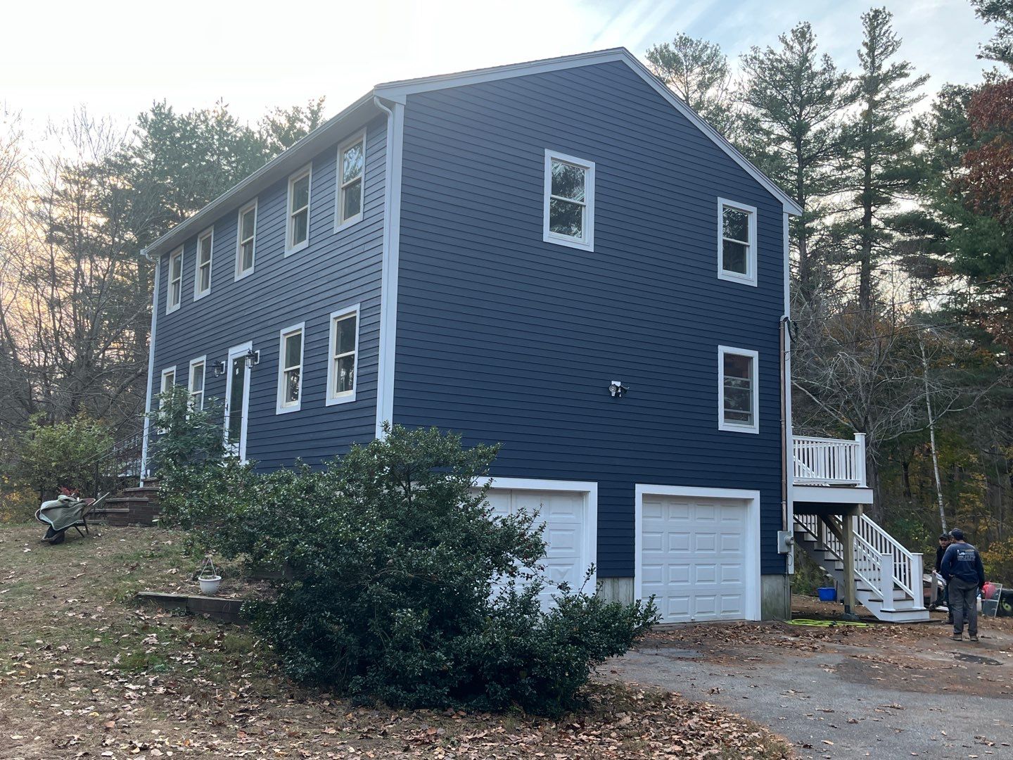 Located in Groveland, MA, this charming two-story blue house features James Hardie Siding and elegant white trim. It boasts a pitched roof, two white garage doors on the ground level, and a side staircase leading up. Surrounded by trees, the scene is complete with someone strolling down the driveway.