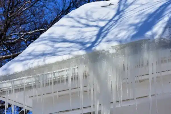 A snow-covered roof with long icicles hanging from the edge hints at potential ice dams that can cause roof damage. The sky is clear and blue, while tree branches cast shadows on the pristine snow below.