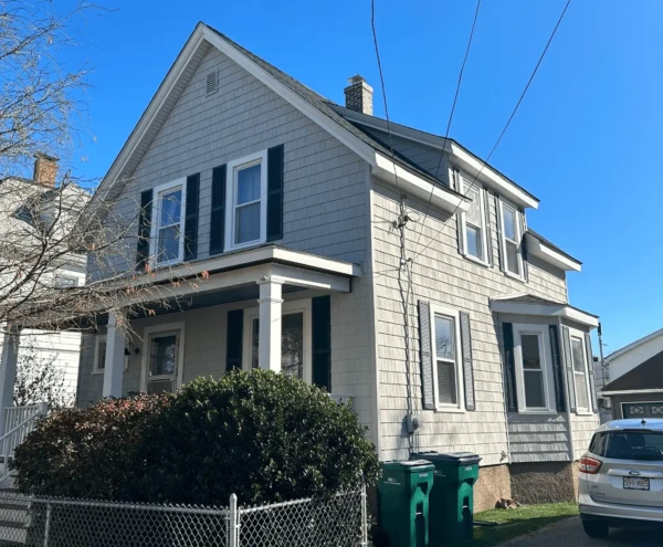 A two-story gray house with dark shutters and a small front porch serves as the ideal setting for window replacement in MA. Bushes adorn the porch while two green recycling bins sit on the driveway. A car is parked nearby, and the sky is clear and blue.