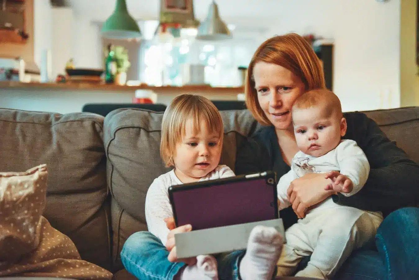 A woman sits on a couch holding a baby, while a toddler next to them explores the tablet, staying connected with family through gentle taps. The softly lit living room provides a warm backdrop, with the kitchen area visible in this intimate scene of togetherness.