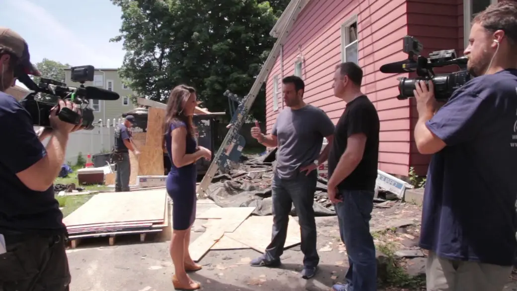 A group of people, including camera operators and journalists, gather at a construction site in Lynn MA beside a pink building on a sunny day. Scattered building materials and debris are visible in the background, capturing the vibrant energy akin to a scene from Flipping Boston.