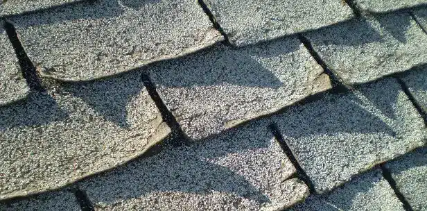 Close-up of a weathered shingle roof showing overlapping, textured shingles with irregular edges, creating a pattern of shadows. The worn surface reveals warning signs that suggest the roof is failing due to prolonged exposure to the elements.