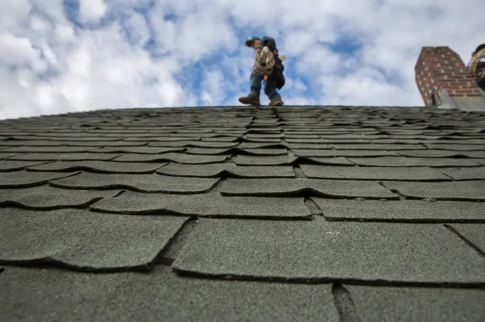 View from a rooftop with damaged, curling shingles in the foreground. Two people with tools and equipment walk along the roof near a brick chimney, preparing for installation of a new roof under a cloudy sky.