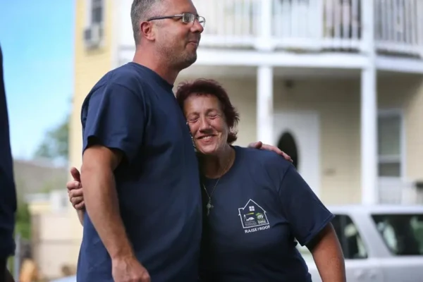 A man and a woman stand outside, both in navy blue shirts. The woman, smiling with her eyes closed, hugs the man—perhaps a homeowner in need—while he looks ahead. A house with visible roof and a car are seen in the background.