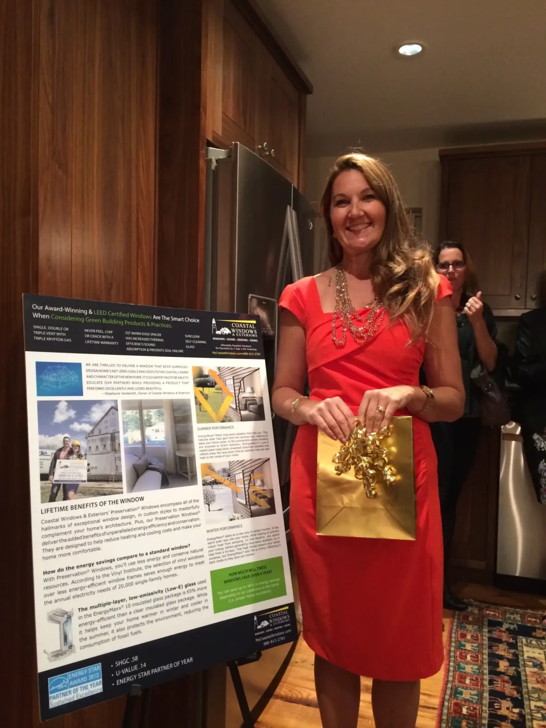 A woman in an orange dress stands next to a presentation board in a kitchen, representing her woman-owned contracting business. She holds a gold gift bag as the board showcases information about energy-efficient windows. People are visible in the background.