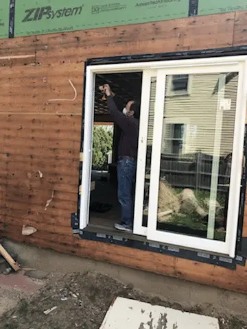 A person is installing a sliding glass door onto a wooden exterior wall of a house, potentially funded by a smart home improvement loan. A residential building looms in the background as they skillfully use a drill while standing halfway through the doorway. Protective covering is partially visible.