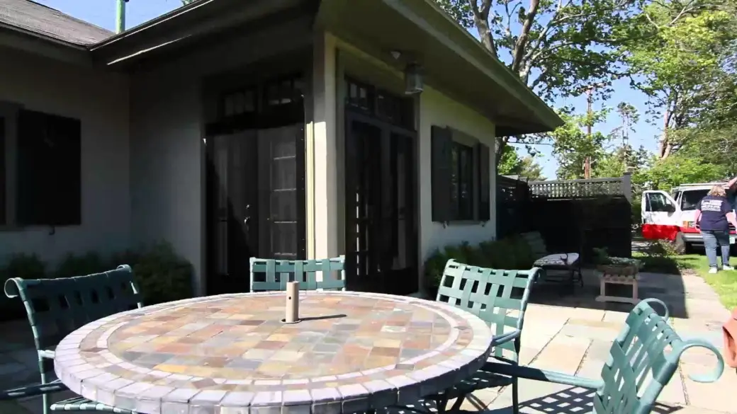 A round patio table with green metal chairs sits on a stone patio outside a house, overlooking a grassy yard and trees—an inviting spot that would earn any Atlantic road testimonial for its relaxing charm. A person stands near a red and white vehicle in the background.