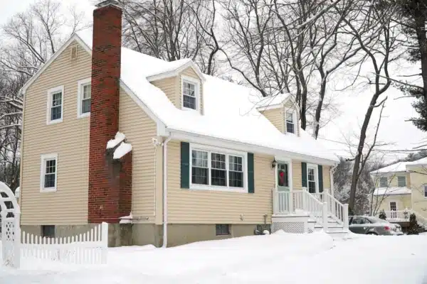 A beige two-story house with replacement windows and a red brick chimney, covered in snow. The house features green shutters and a front porch adorned with a wreath on the door. Leafless trees and another snow-covered house are visible in the background.