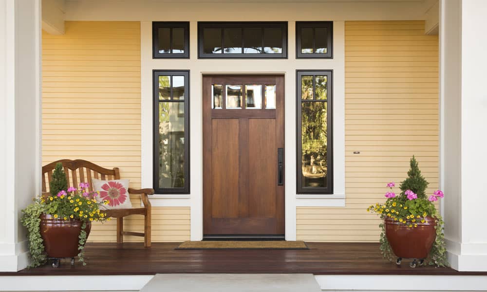A welcoming front porch with a wooden door flanked by sidelights. The yellow siding is complemented by large potted plants with vibrant flowers. A wooden bench with a floral cushion is on the left, all on dark wooden flooring.