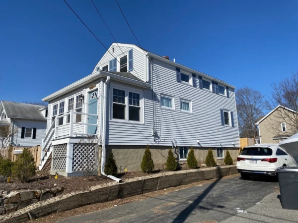 A two-story white house with fire-resistant vinyl siding features multiple windows and a small porch. Its surrounded by shrubs and a stone retaining wall. A parked white SUV is visible in the driveway. The sky is clear and blue, completing the serene scene.