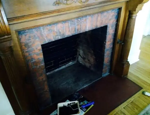 An empty fireplace with a wooden mantel and brick interior sits ready for a DIY fireplace project. Cleaning supplies, including a dustpan and brush, are placed on the floor in front of this budget renovation scene.