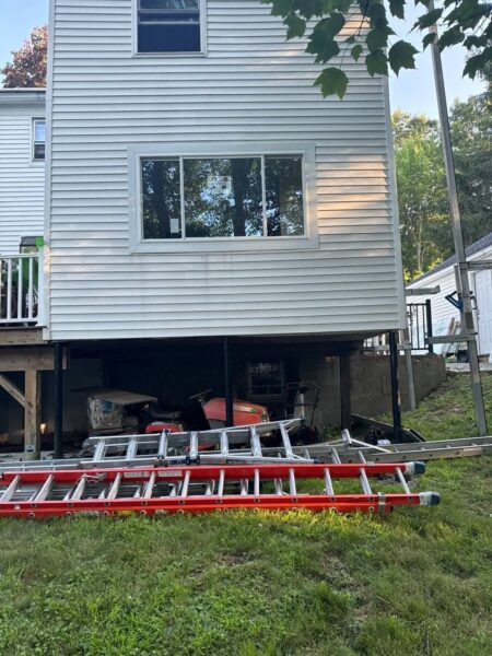 A house with white siding appears elevated on one side, revealing an open space underneath. Below the house, there are two ladders lying on the grass, along with various equipment and tools. Trees and a wooden fence are visible in the background.