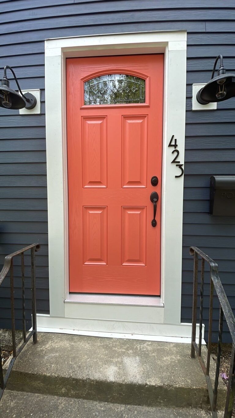 A coral-colored door with a black handle is centered on a dark blue house with white trim. The house number 425 is mounted vertically on the right. Two black wall lanterns flank the door. Steps lead up to the entrance.