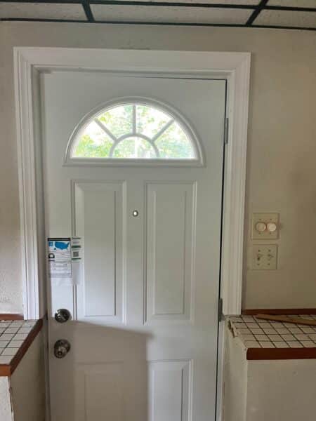 The front door installation in Swampscott, MA, features a white door with a crescent window and small peephole. A label is attached to the left side, while on the right, a wall switch with two dials is visible against an off-white wall with tiled half-wall sections.