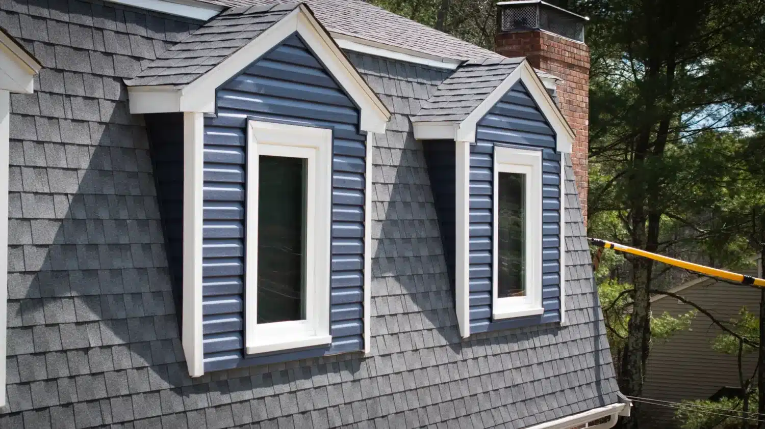 Rooftop with gray shingles and two dormer windows featuring blue siding and white trim, sparking a common debate of whether to repair or replace the roof. A brick chimney stands on the right, framed by green trees in the background.