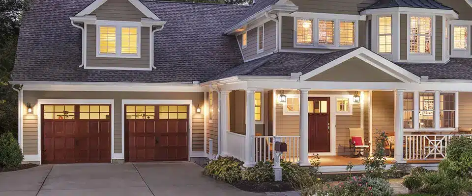 A cozy two-story house with warm lights on inside, featuring a front porch with white railings, a red door, large windows and quality garage doors. New roofing and energy-efficient windows & doors may qualify for a tax credit. Shrubs and a driveway are in front.