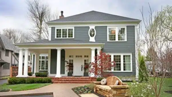 Two-story gray house with white trim and a covered porch. The front yard features a stone pathway, manicured shrubs, and a small tree with red leaves. Large windows, acclaimed in window contractor reviews, and a circular window accent adorn the exterior. Cloudy sky background.