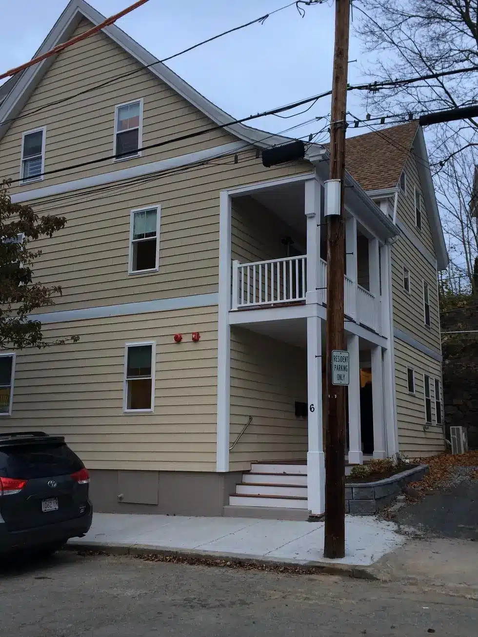 A beige three-story Habitat for Humanity-North Shore house with white trim, a covered porch, and stairs leading to the entrance. A car is parked in front, near a utility pole and sign. Leafless trees stand in the background.