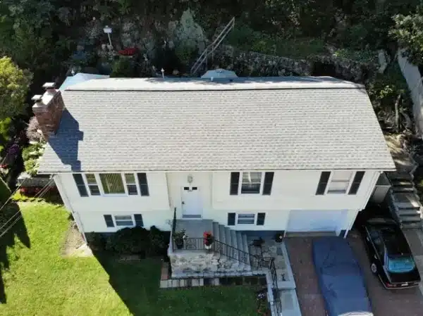 Aerial view of a two-story white house with a gray roof, showcasing expert roofing in Hanover, MA. The home features a front entrance with stairs, a small porch, and a driveway where a car rests under a tarp. Lush green lawn and towering trees complete the serene setting.