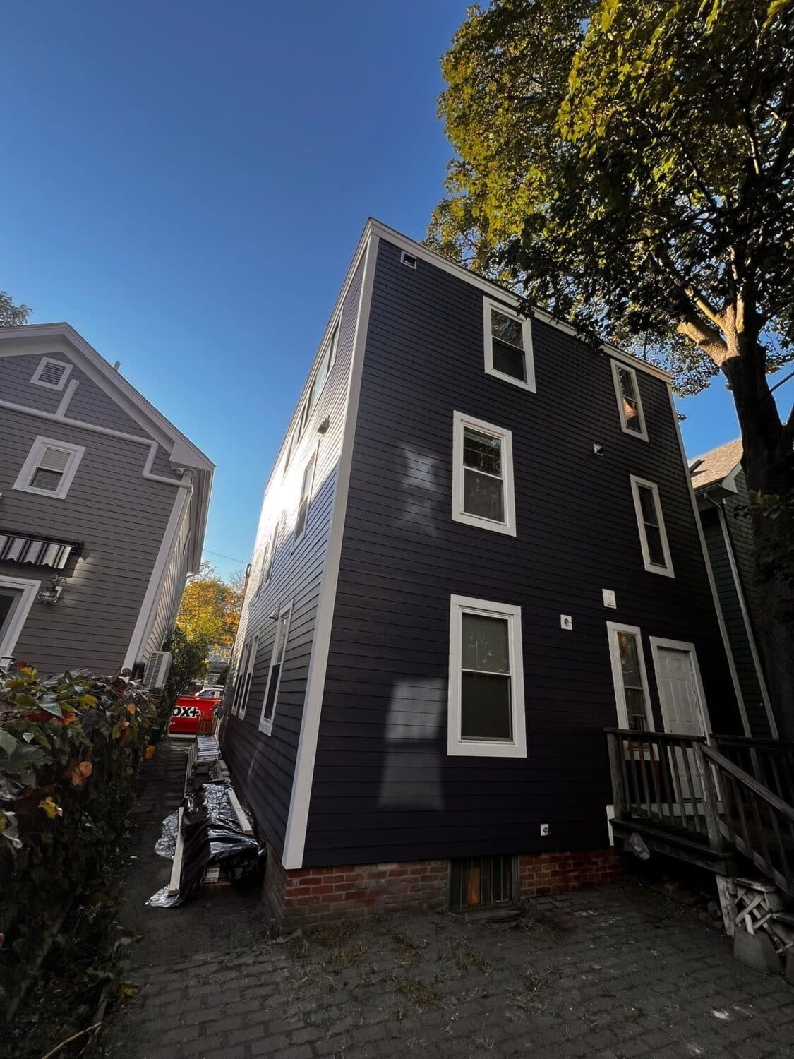 A narrow, three-story dark gray house with white trim stands against a clear blue sky. Sunlight illuminates one side. The house is flanked by trees and another house. Theres a small porch and a visible red vehicle in the background.