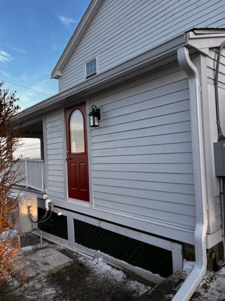 A house with white siding and a red door elevated above ground level. Theres a white gutter system and a lantern-style light fixture beside the door. The sky is clear, and theres a bush with orange-tinted branches.