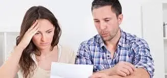 A worried couple sits together, examining a roof inspection checklist. The woman has a hand on her forehead, expressing concern, while the man looks intently at the paper. They both seem focused and slightly stressed.