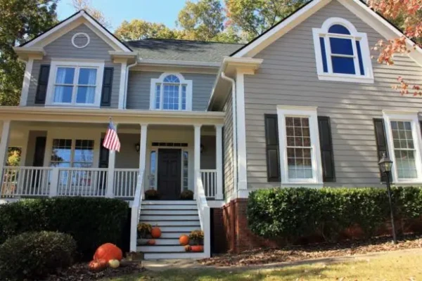 A two-story house with gray siding and white trim boasts a welcoming front porch adorned with pumpkins. An American flag flutters proudly, while green shrubs and trees with autumn leaves complete the scene—ideal inspiration for your next fall home improvement project.