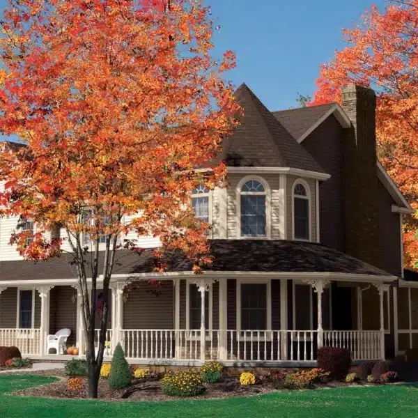 A two-story Victorian-style house with a wraparound porch is nestled among vibrant autumn trees, their orange and red leaves whispering energy savings tips. Under the clear blue sky, the neatly manicured lawn features small hedges and flowers, completing this picturesque and efficient setting.