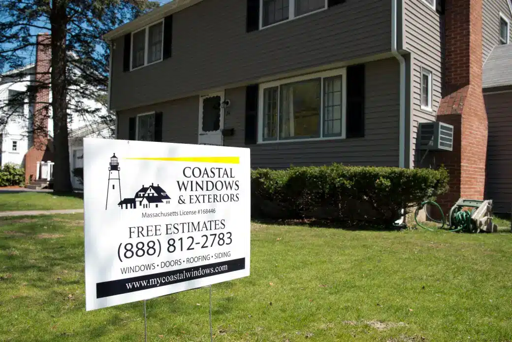 A sign in front of a house reads Coastal Windows & Exteriors and offers free estimates for windows, doors, roofing, and replacing your home siding. Both the house and lawn are visible in the background, along with a phone number and website.