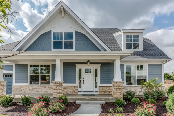 Charming two-story house with blue vinyl siding and stone accents, maintaining a classic look. The gabled roof and white front door are centered under a covered porch supported by square columns. Bushes and blooming flowers line the walkway, framed by a bright, partly cloudy sky overhead.