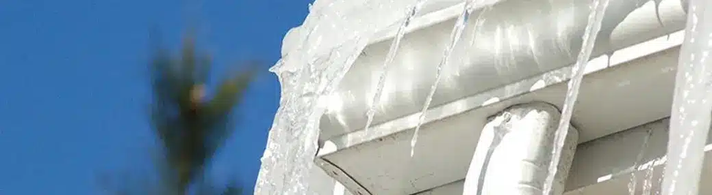 Icicles hang precariously from a snow-covered roof, hinting at potential roof ice dams against the clear blue sky. A blurred tree is visible in the background, adding a touch of nature to the serene winter scene.