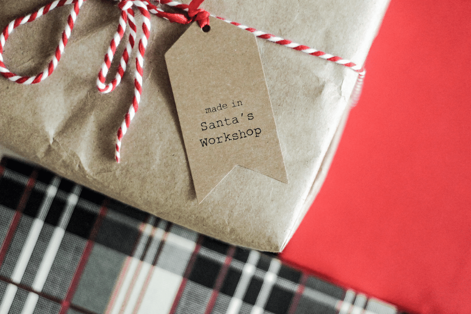 A close-up of a gift wrapped in brown paper with red and white twine, featuring a tag that reads made in Santa’s Workshop. The present sits on a red and plaid patterned surface, ready for delivery down the chimney this holiday season.