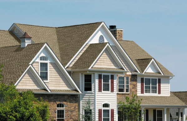 A suburban house with a complex GAF roofing design features brown shingles, stone and beige siding, and several white-framed windows with red shutters. The sky is clear, and there are leafy green trees in the foreground.