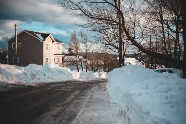 A residential street is lined with large piles of snow. A two-story house on the left, part of a winter home maintenance checklist, stands beside leafless trees stretching into a partly cloudy sky. The road is wet but clear of snow, capturing a quiet winter scene.