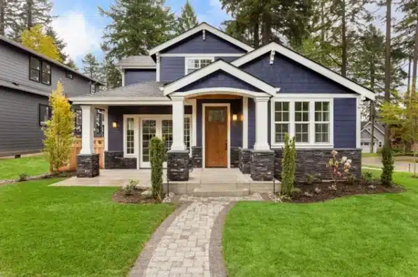 A charming two-story house with navy blue siding, white trim, and a wooden front door boasts large windows that highlight the benefits of replacing windows. It features a stone-accented porch and a paved walkway leading through a well-manicured lawn, surrounded by trees.