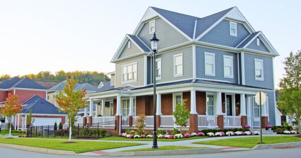 A large two-story house with gray siding and brick lower walls sits on a corner lot, showcasing some of the best home improvements like a wraparound porch, manicured lawns, and white railing. A lamppost and stop sign are in front, with a tree-lined hill as the backdrop.