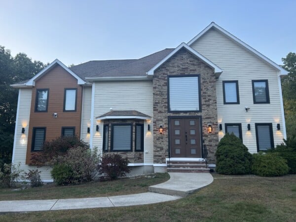 Two-story house with a mix of beige James Hardie siding, stone, and wood accents. The front features a large brown door with sidelights, multiple windows, and wall lights. A curved pathway leads from the entrance, bordered by shrubs and a lawn in Methuen MA.