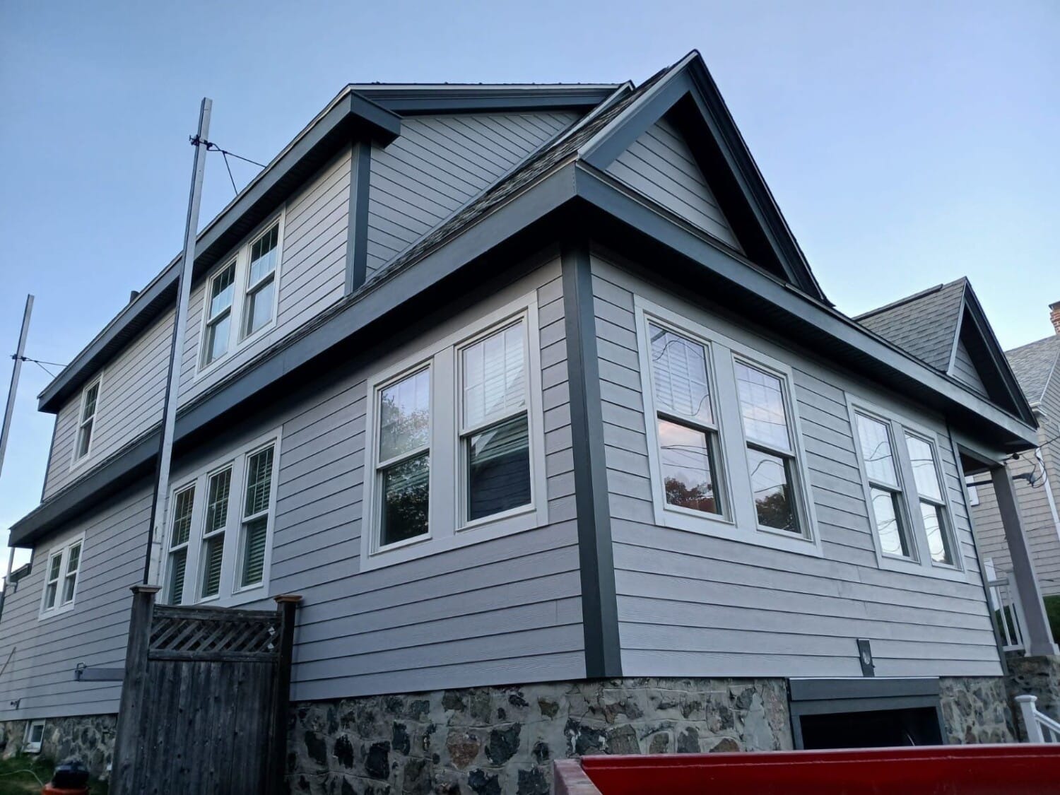 The two-story house in Swampscott, MA, showcases elegant gray James Hardie siding with a stone foundation. White-framed windows complement the roofs mix of light and dark gray shingles. Under a clear sky, a wooden gate is visible on the left.