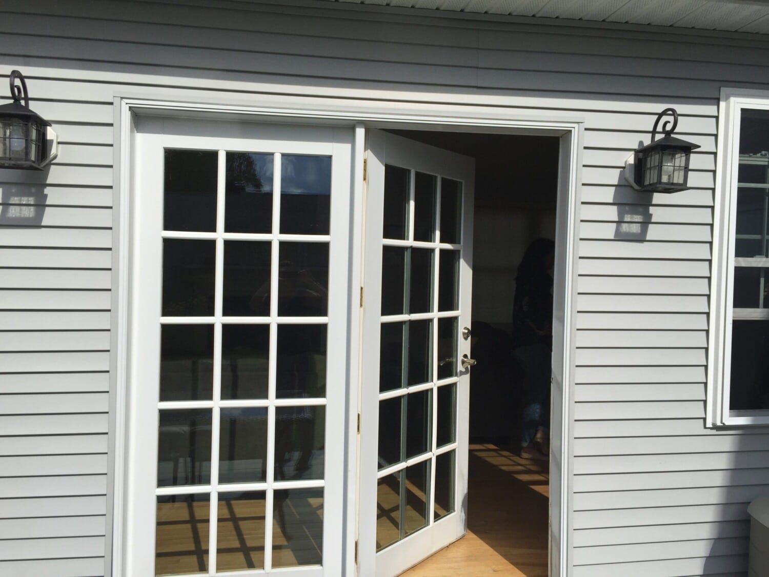 A set of white French doors, one open, leads from the outside into a house with gray siding. Reminiscent of a sliding glass door, two black wall lanterns flank the doors as sunlight streams onto a wooden floor inside.