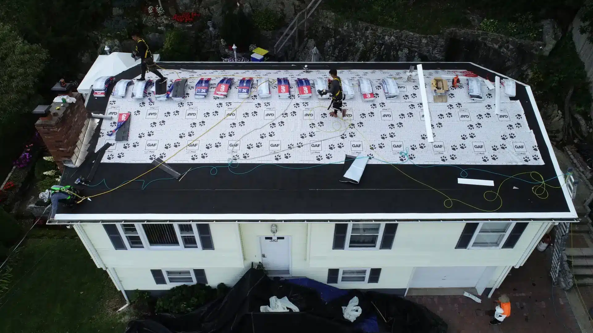 Aerial view of workers installing a new roof on a two-story house. Roofing materials and tools are spread out, with safety ropes secured, as New England roofing techniques are applied to the roof now partially covered with underlayment and shingles.