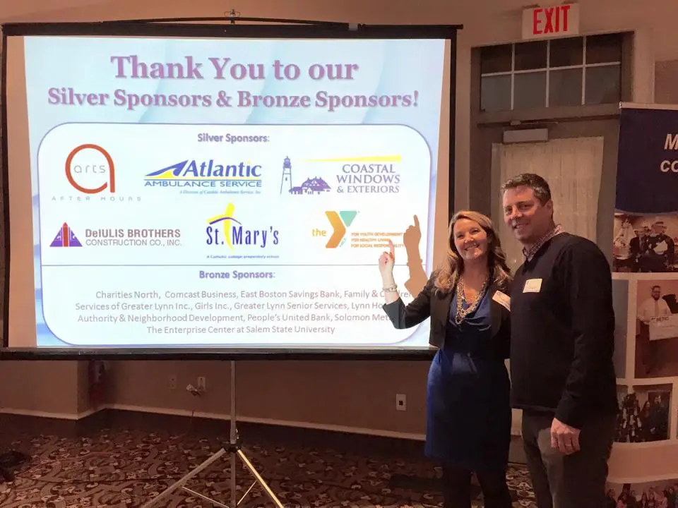 Two people stand smiling in front of a projection screen displaying a thank you message to silver and bronze sponsors, highlighting the community impact made possible by the support of multiple companies whose logos and names are listed.