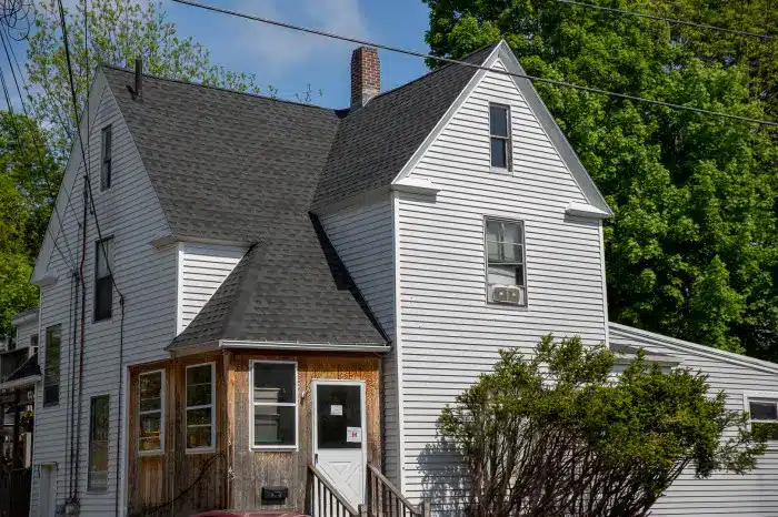 A two-story white house with a steep black roof and a wooden front porch stands proudly amidst green trees and bushes. A chimney graces the roof, newly installed and glistening under the blue sky. Overhead power lines add to the timeless charm of this picturesque scene.