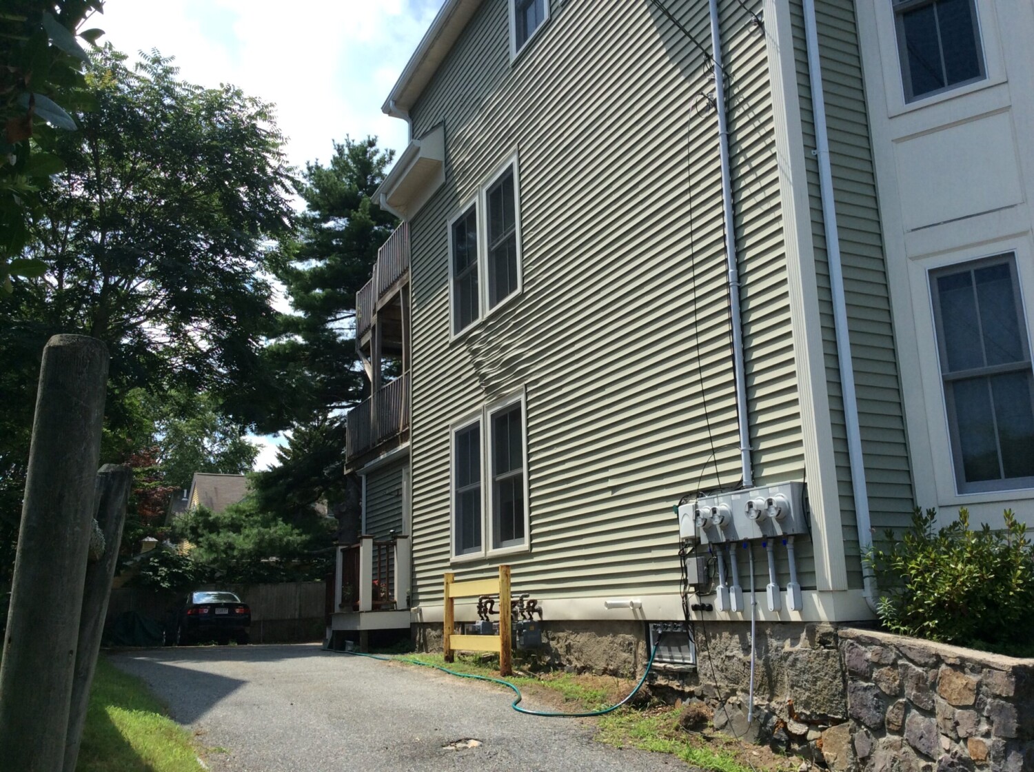 A narrow driveway runs alongside a light-colored, three-story house with vinyl siding and multiple windows. Utility meters and a garden hose are mounted near the stone foundation. Trees, a parked car, and signs of melted vinyl siding repair are visible in the background.