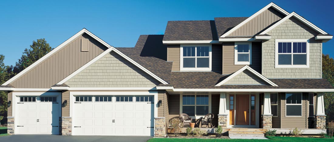 A two-story suburban house with a double garage, gray siding, and stone accents. The home features large windows, a front porch with seating, and a well-maintained lawn against a clear blue sky.