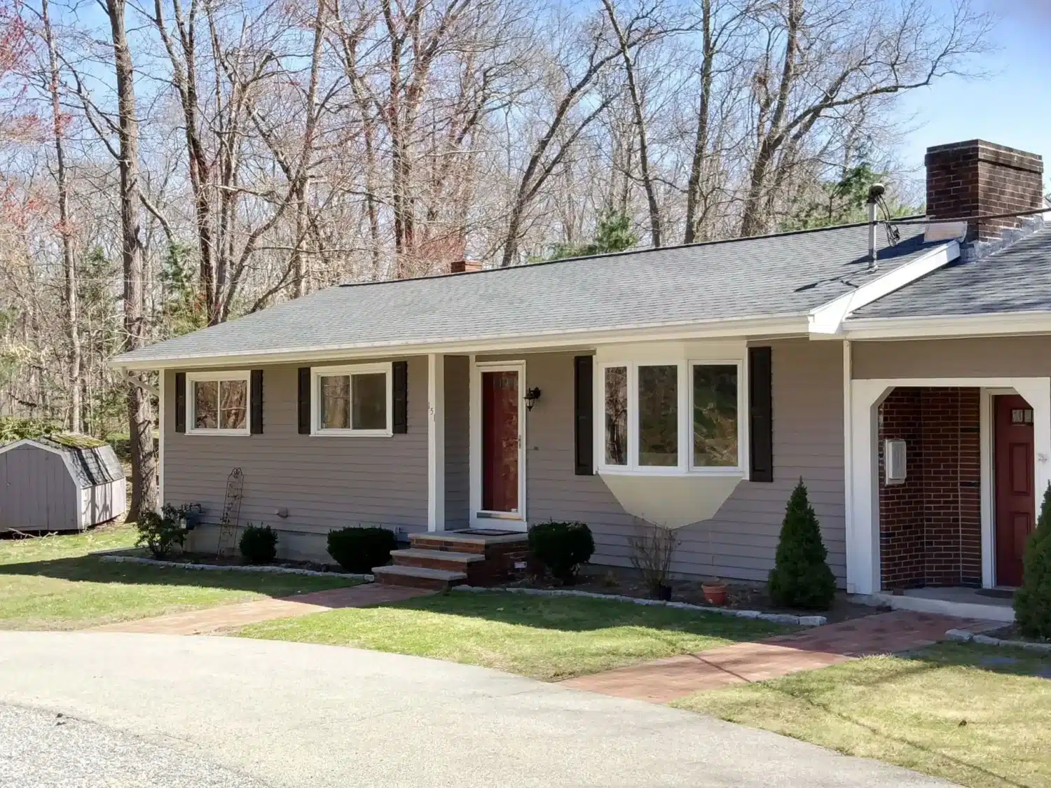 A single-story house with sustainable roofing, light brown siding, a red door, and a brick chimney. It features white-framed windows and a small front garden. A shed sits in the background, surrounded by bare trees under a clear sky.