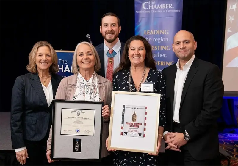 Five people stand together smiling, two holding framed awards, at the North Shore Chamber Salute to Veterans Breakfast. The event is organized by The North Shore Chamber of Commerce. A banner in the background reads Leading the region. Investing together. Empowering success.