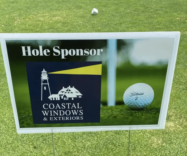 Sign on grass for Coastal Windows & Exteriors as hole sponsor, featuring an illustrated lighthouse and house. At a North Shore Chamber golf outing, a close-up of a golf ball by the hole captures the spirit of sponsorship.