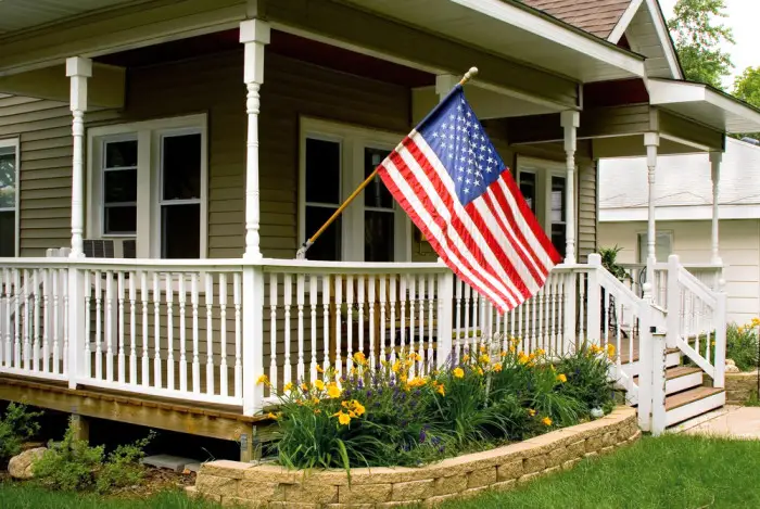 A porch with white railings and columns displays an American flag, adding curb appeal. Yellow flowers and green plants grow in a landscaped bed in front of the beige house exterior with white trim.