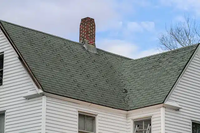 A close-up view of a house’s gray shingled roof with a brick chimney highlights the importance of winter roof repair. The white siding and windows are visible, set against a mostly cloudy sky with patches of blue.